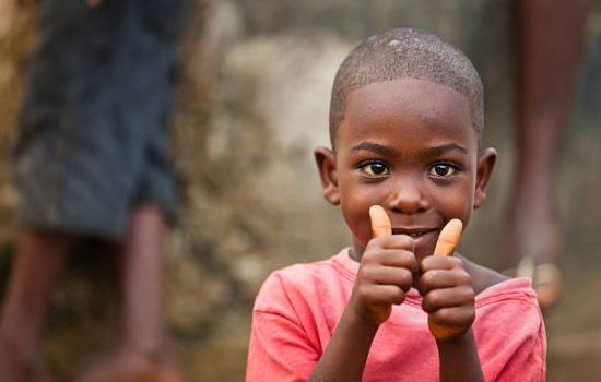 A young African boy sitting near a public water pump smiling white giving a thumbs up.